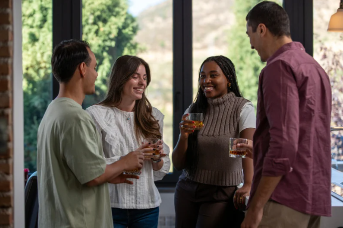 rozmowa z alkoholikiem; Na zdjęciu widać cztery dorosłe osoby stojące razem w jasnym pomieszczeniu, trzymające w dłoniach szklanki z alkoholem. W kontekście tematu można to opisać jako rozmawiać z alkoholikiem – spotkanie, podczas którego jedna z osób potrzebuje pomocy, a pozostali próbują ją wesprzeć rozmową. Jej przebieg jest spokojny i oparty na słuchaniu, bez widocznych oznak konfliktu. Faktycznie widoczne są napoje alkoholowe, co może symbolizować problem nadużywania alkoholu i moment, w którym dana osoba zaczyna odczuwać konsekwencje nadużywania alkoholu