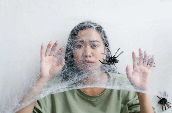 czym jest fobia - Na zdjęciu widać osobę stojącą za gęstą pajęczyną, z wyraźnym napięciem i niepokojem na twarzy, gdy tuż przed nią znajduje się pająk. Jej gesty sugerują chęć odsunięcia zagrożenia, ale jednocześnie bezradność. Taka scena pokazuje, jak lęk znacząco wpływa na emocje i zachowanie człowieka, ograniczając swobodę ruchu i poczucie kontroli. Może to być fobia związana z pająkami, czyli silna, irracjonalna reakcja, która pojawia się automatycznie. Cały obraz dobrze oddaje intensywność lęku