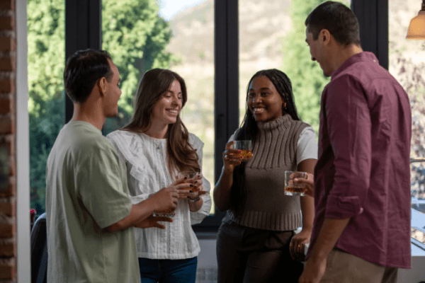 rozmowa z alkoholikiem; Na zdjęciu widać cztery dorosłe osoby stojące razem w jasnym pomieszczeniu, trzymające w dłoniach szklanki z alkoholem. W kontekście tematu można to opisać jako rozmawiać z alkoholikiem – spotkanie, podczas którego jedna z osób potrzebuje pomocy, a pozostali próbują ją wesprzeć rozmową. Jej przebieg jest spokojny i oparty na słuchaniu, bez widocznych oznak konfliktu. Faktycznie widoczne są napoje alkoholowe, co może symbolizować problem nadużywania alkoholu i moment, w którym dana osoba zaczyna odczuwać konsekwencje nadużywania alkoholu
