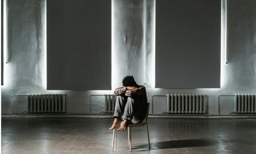 stany depresyjne a depresja - zdjęcie przedstawia mężczyznę, który siedzi w dużym ponurym pomieszczeniu, skulony, chowa twarz w ramionach