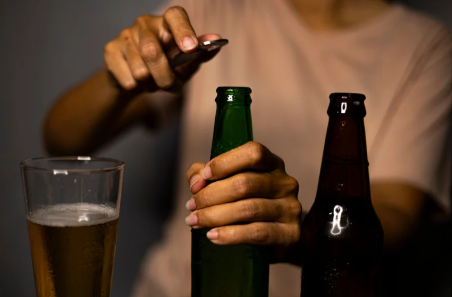 drżenie rąk po alkoholu - zdjęcie przedstawia rękę mężczyzna który w jednym ręku trzyma piwa a w drugiej otwieracz, obok stoi szklanka pełna piwa i drugie otwarte piwo